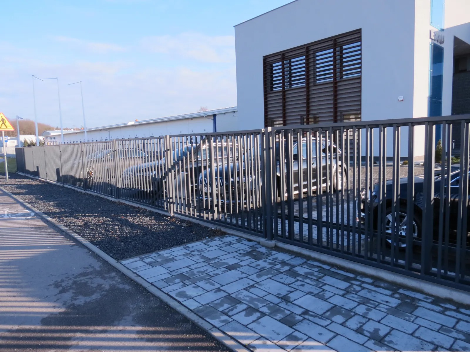 Nowoczesne ogrodzenie pionowe z metalowych prętów na tle budynku biurowego i zaparkowanych samochodów.