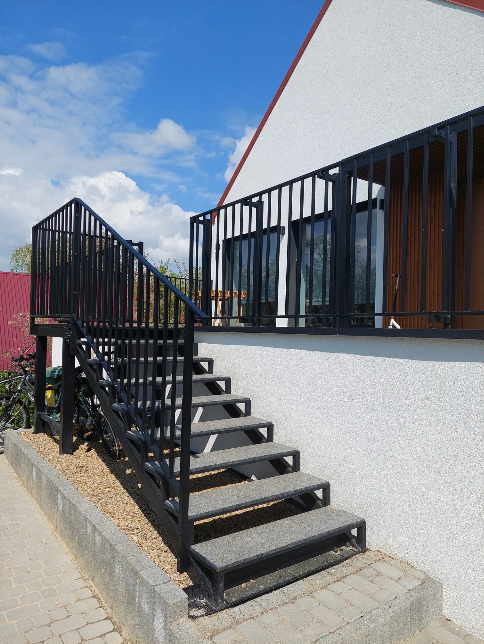 Zewnętrzne schody metalowe z szarymi stopniami prowadzące na balkon z czarną balustradą, obok zaparkowany rower.