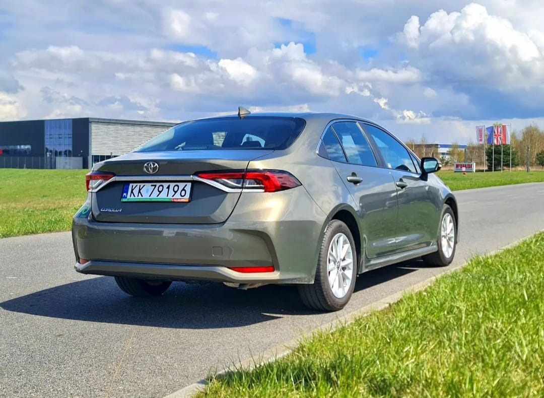 Szary sedan Toyota Corolla, widok z tyłu, zaparkowany na poboczu drogi z zieloną trawą, nowoczesny budynek w tle pod błękitnym niebem z chmurami.