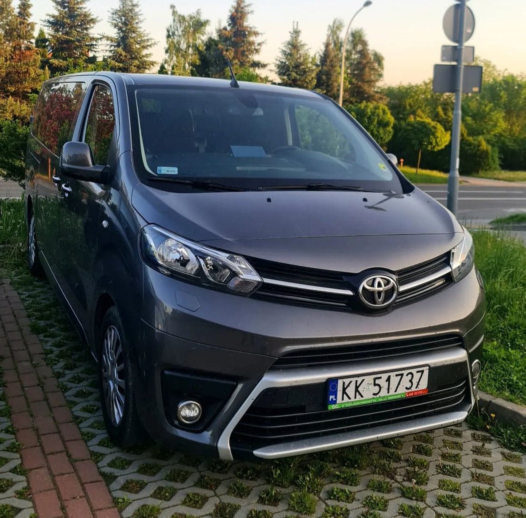 Szary minivan Toyota Proace Verso z polską tablicą rejestracyjną KK 51737, zaparkowany na kostce brukowej z zieloną trawą, z drzewami w tle.