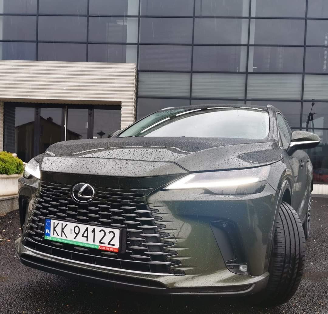 Przód ciemnozielonego samochodu Lexus NX z polską tablicą rejestracyjną KK 94122, zaparkowanego przed nowoczesnym budynkiem z szklaną fasadą w deszczowy dzień.