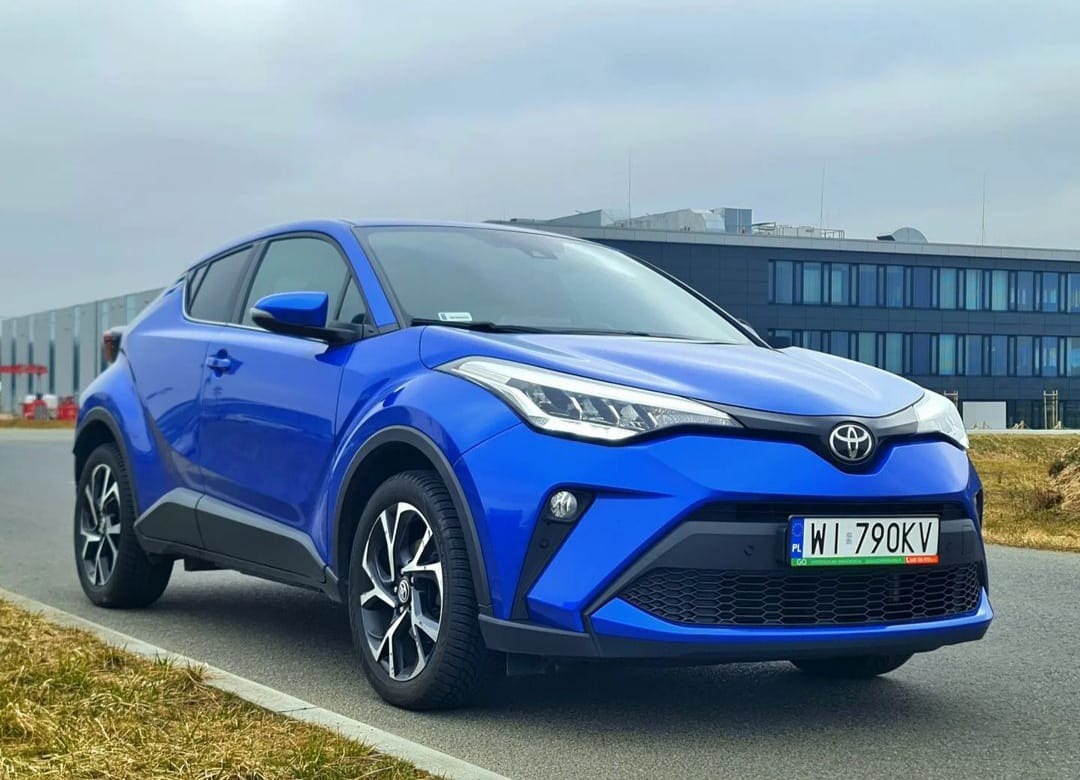Niebieski samochód Toyota C-HR zaparkowany na asfalcie z budynkiem w tle, widoczna tablica rejestracyjna WI 790KV.