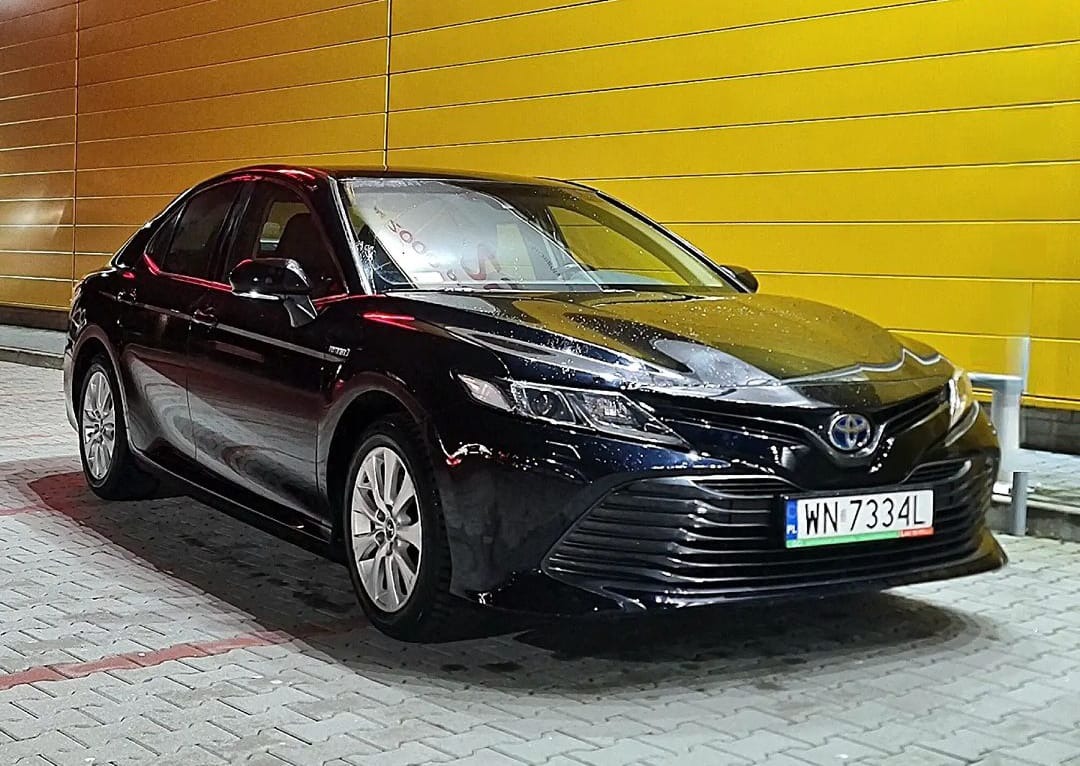 Błyszczący, czarny sedan marki Toyota Camry, zaparkowany na kostce brukowej z żółtym, metalicznym tłem. Widoczne detale: aluminiowe felgi, reflektory LED, tablica rejestracyjna WN 7334L.