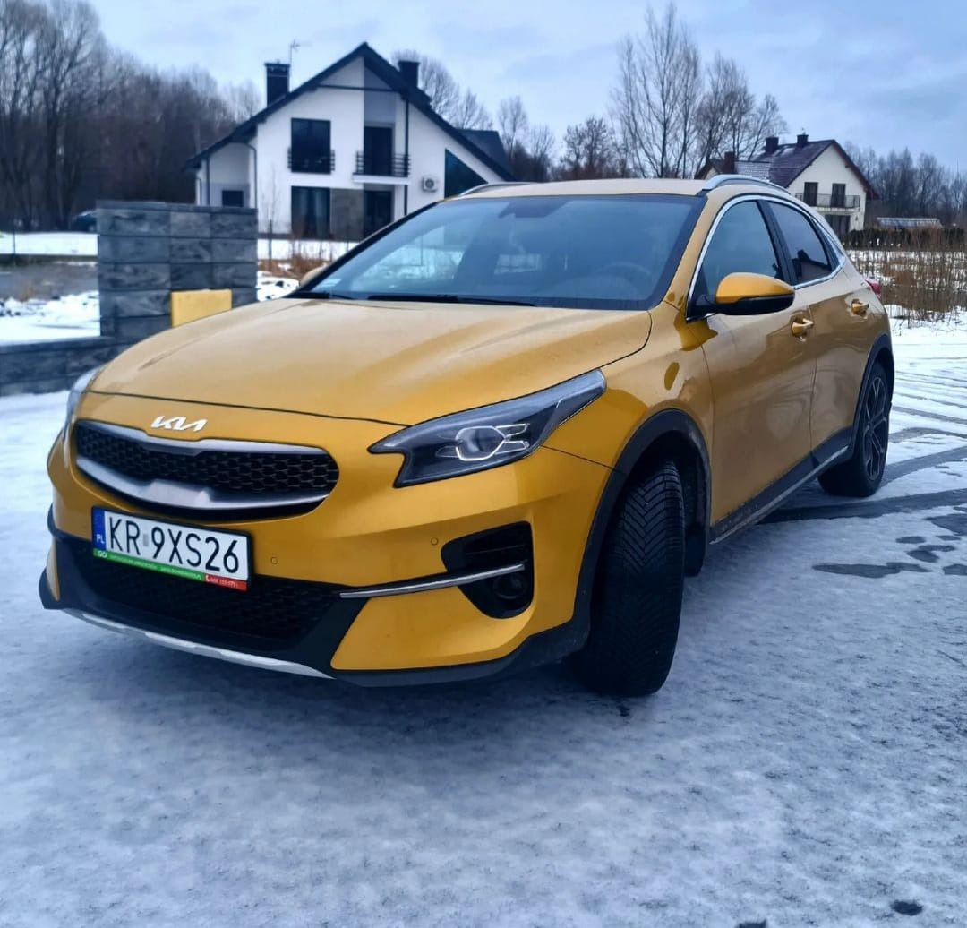 Żółty samochód Kia XCeed z numerem rejestracyjnym KR 9XS26 zaparkowany na śniegu przed domem.