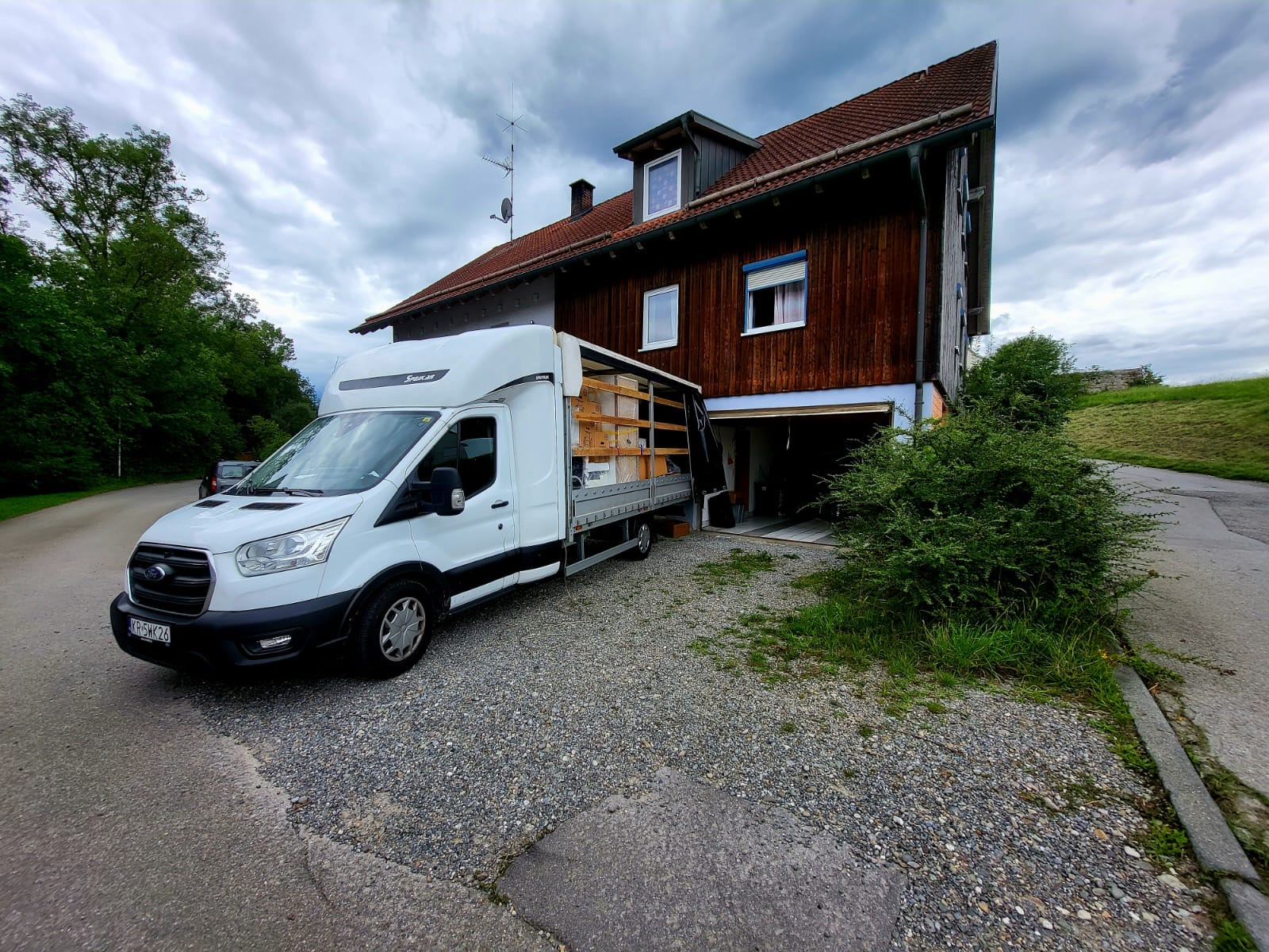 Biały samochód dostawczy z ładunkiem stoi na żwirowym podjeździe przed drewnianym domem w okolicy Krakowa, sugerując transport mebli lub innych przedmiotów.