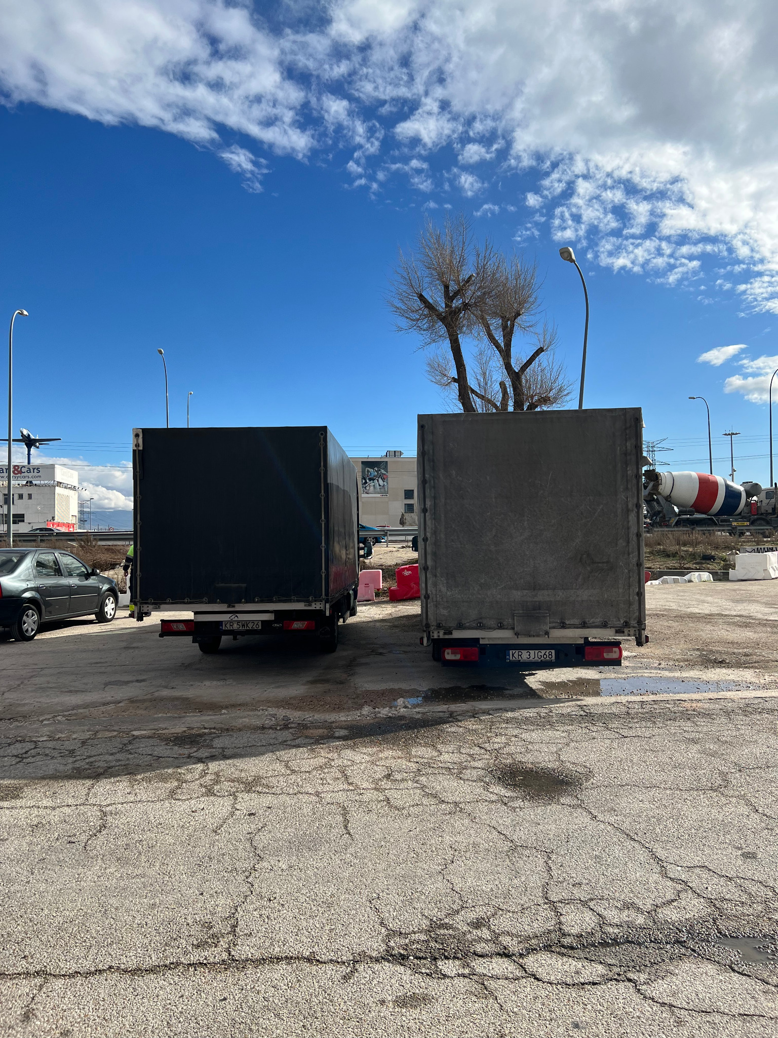 Dwa busy z plandekami zaparkowane na placu, widok z tyłu, w tle budynki i mikser do betonu.