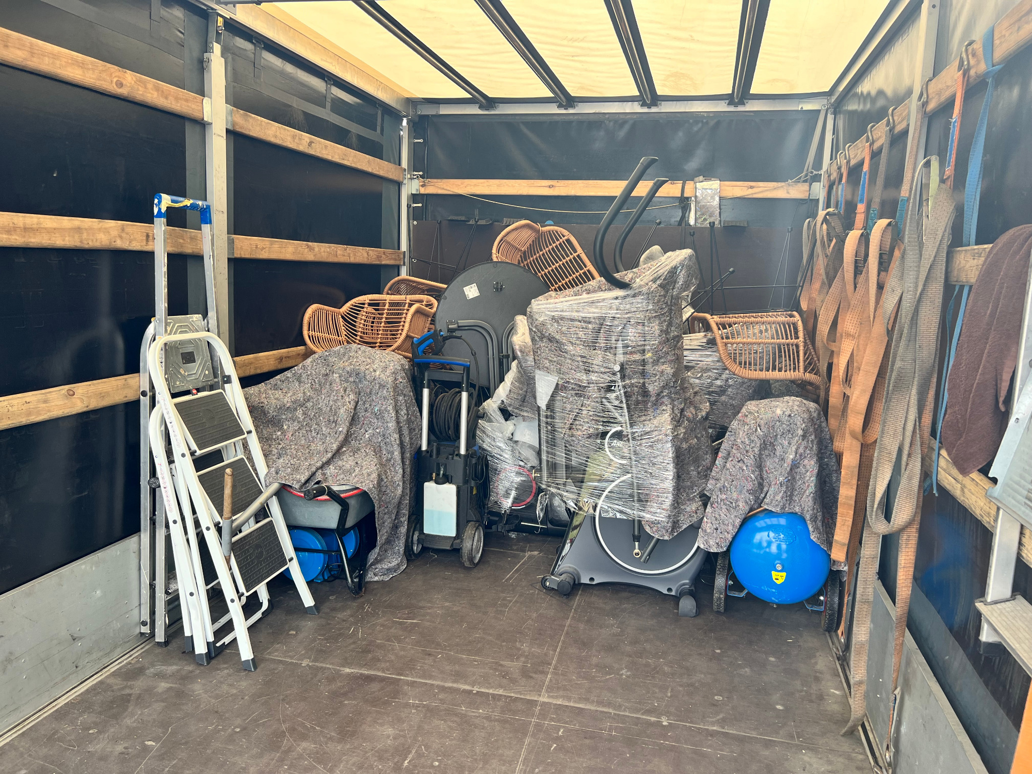 Wnętrze skrzyni ładunkowej wypełnione meblami rattanowymi, sprzętem do czyszczenia i ćwiczeń, zabezpieczonymi folią i kocami, z drabinką i pasami mocującymi po bokach.