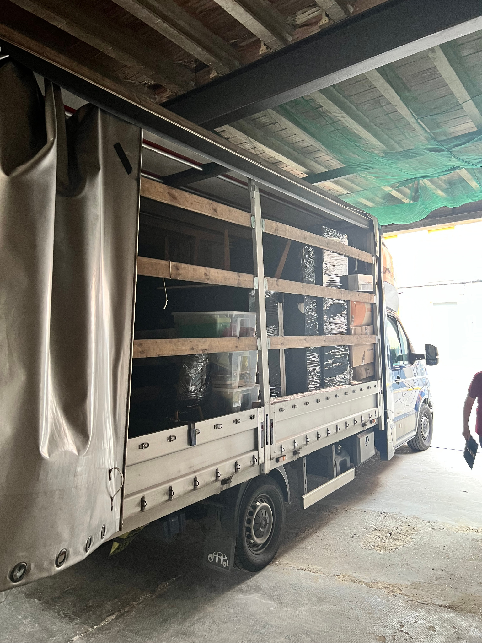 Ciężarówka z plandeką, częściowo załadowana kartonami i plastikowymi pojemnikami, widoczna w magazynie z drewnianym stropem i zieloną siatką zabezpieczającą.