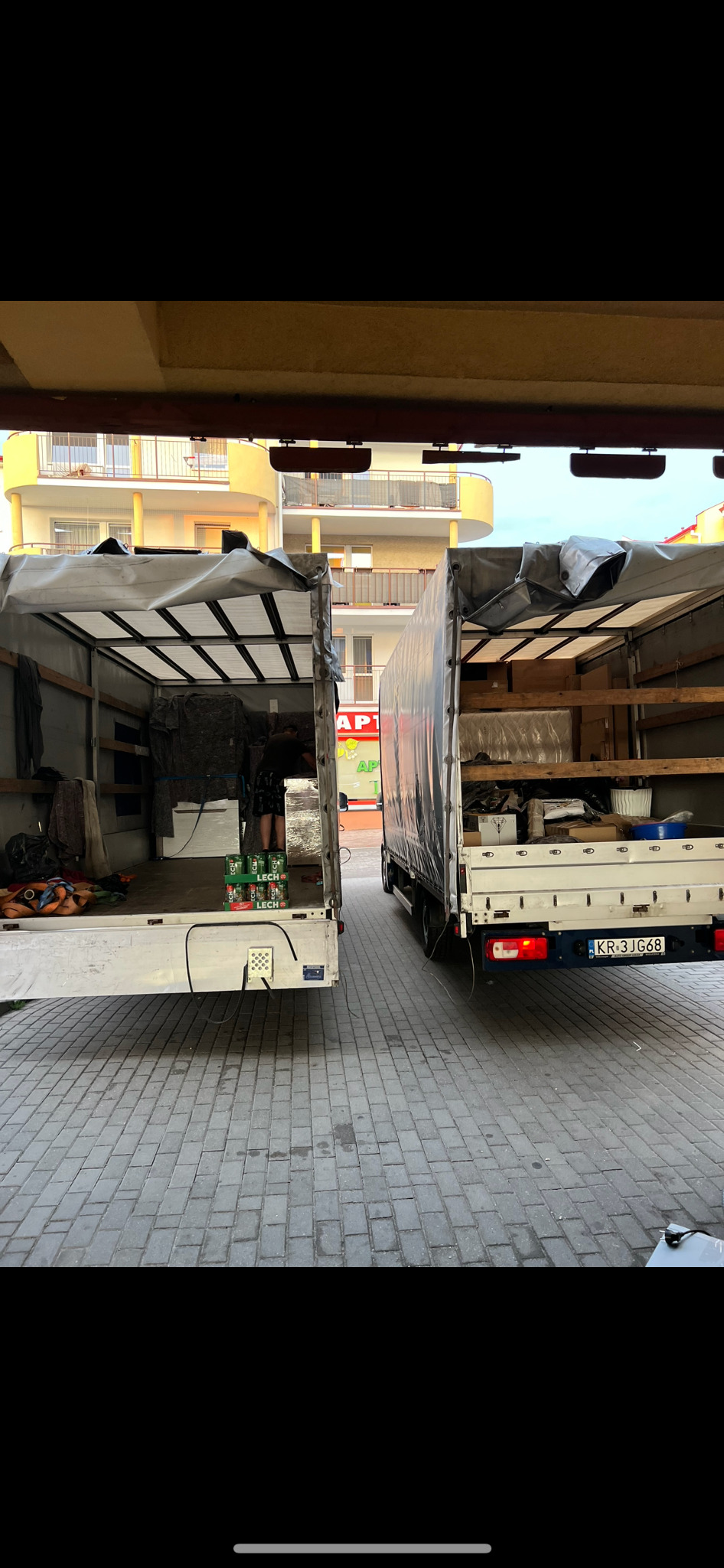 Dwa busy z otwartymi przestrzeniami ładunkowymi, jeden z załadowanym sprzętem i napojami, drugi z częściowo załadowanymi kartonami i meblami, zaparkowane na brukowanym placu przed budynkiem.