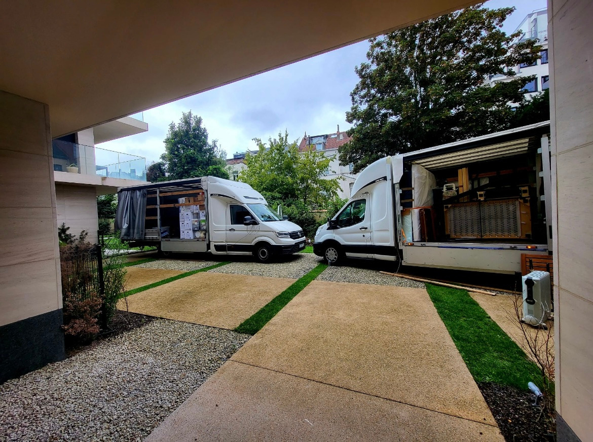 Dwa białe busy dostawcze zaparkowane na posesji z nowoczesnym budynkiem w tle, jeden z otwartą przestrzenią ładunkową wypełnioną meblami i kartonami.