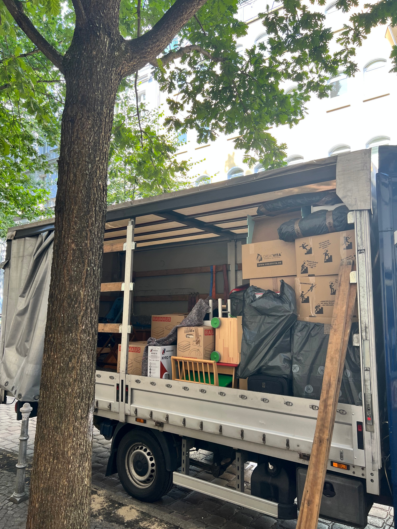 Ciężarówka do przeprowadzek załadowana kartonami, meblami i workami, zaparkowana przy chodniku z drzewem w tle.