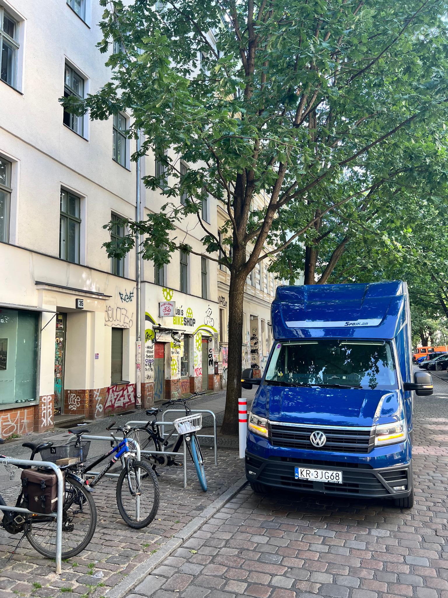 Niebieski samochód dostawczy marki Volkswagen Spojkar zaparkowany na brukowanej ulicy w pobliżu budynku z graffiti i drzewami.