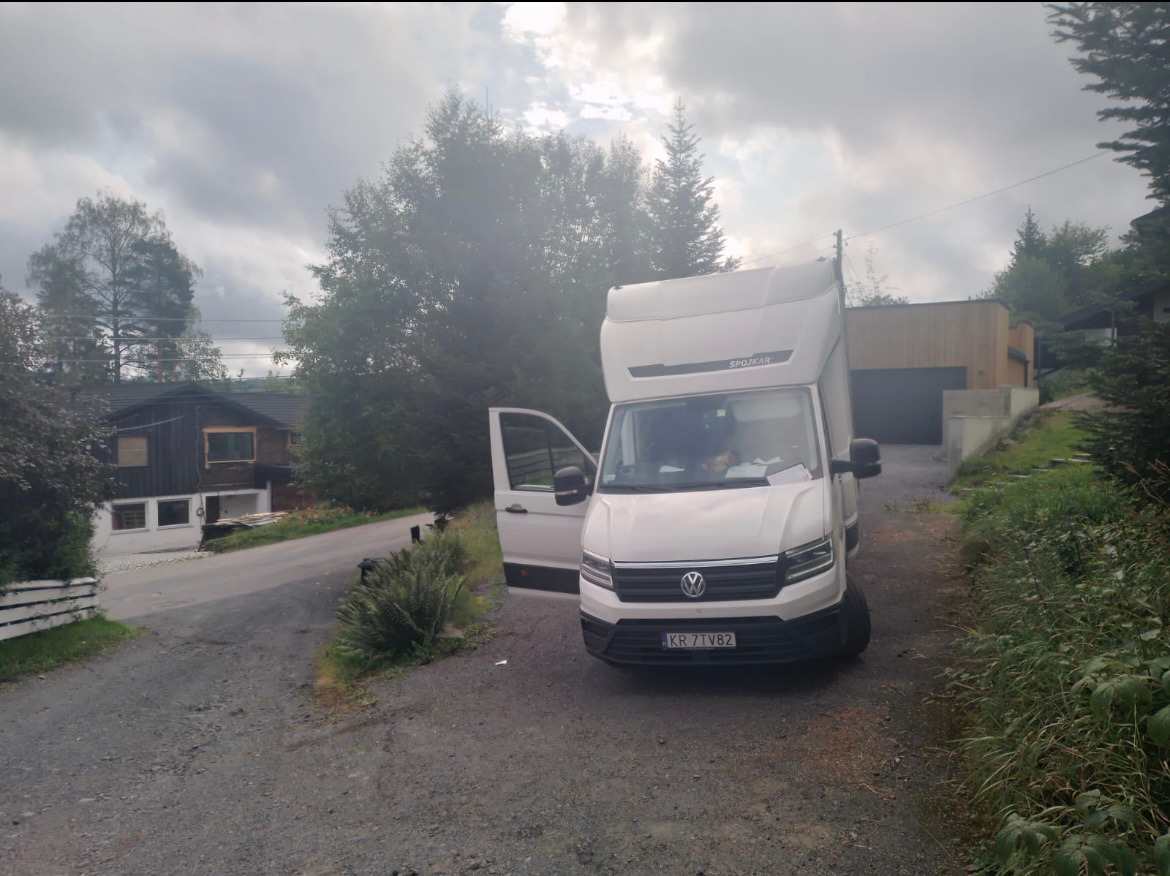 Biały bus Volkswagen Crafter z otwartymi drzwiami kierowcy zaparkowany na żwirowej drodze w górzystym terenie, widoczne domy w tle.