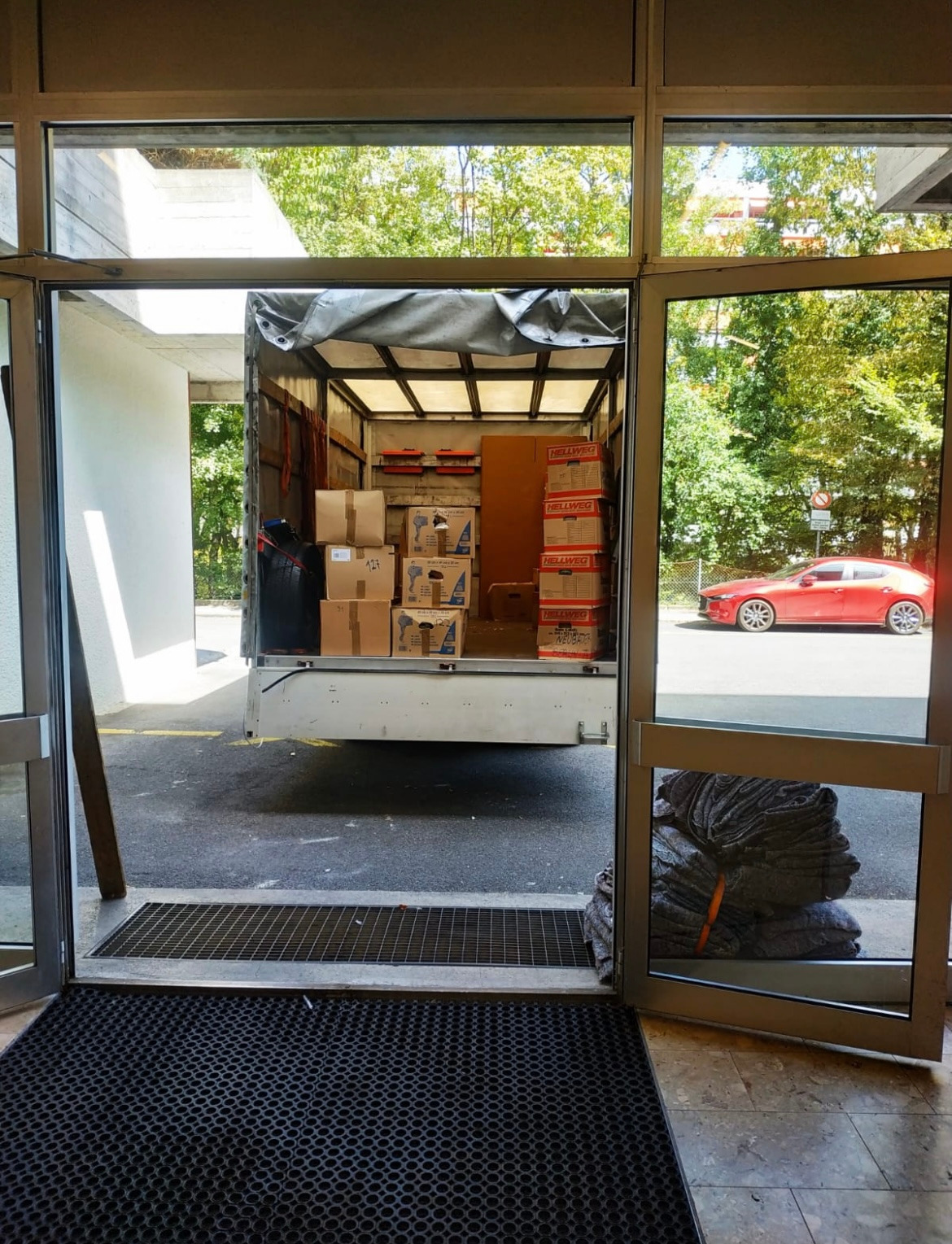 Widok z wnętrza budynku na otwartą pakę ciężarówki załadowaną kartonami i narzędziami, przez otwarte drzwi widoczny czerwony samochód na zewnątrz.
