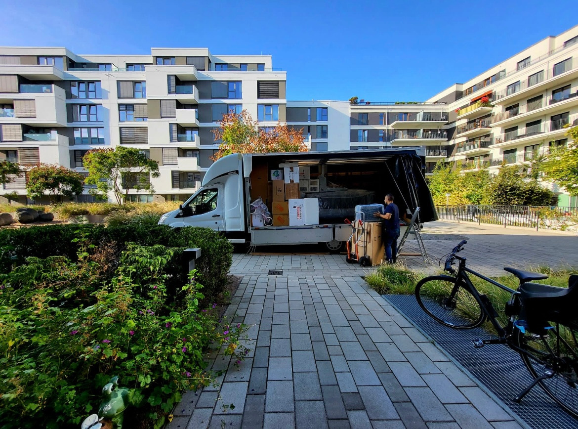 Biała furgonetka z otwartą przestrzenią ładunkową wypełnioną kartonami i meblami na tle nowoczesnych budynków mieszkalnych, pracownik transportuje zapakowane przedmioty, obok stoi rower.