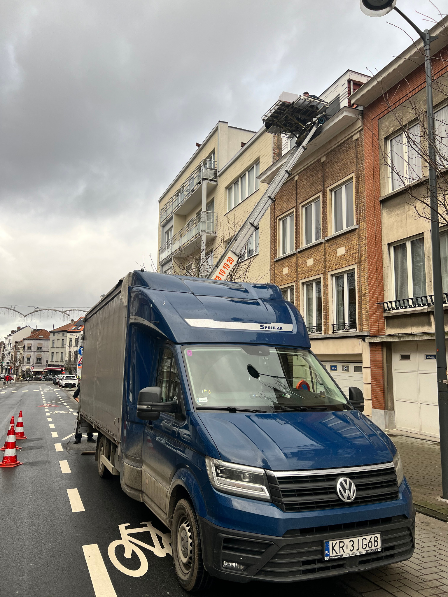 Niebieski samochód dostawczy marki Volkswagen z podnośnikiem meblowym, ustawiony na ulicy z wyznaczonym pasem dla rowerów, podczas załadunku do budynku mieszkalnego w pochmurny dzień.