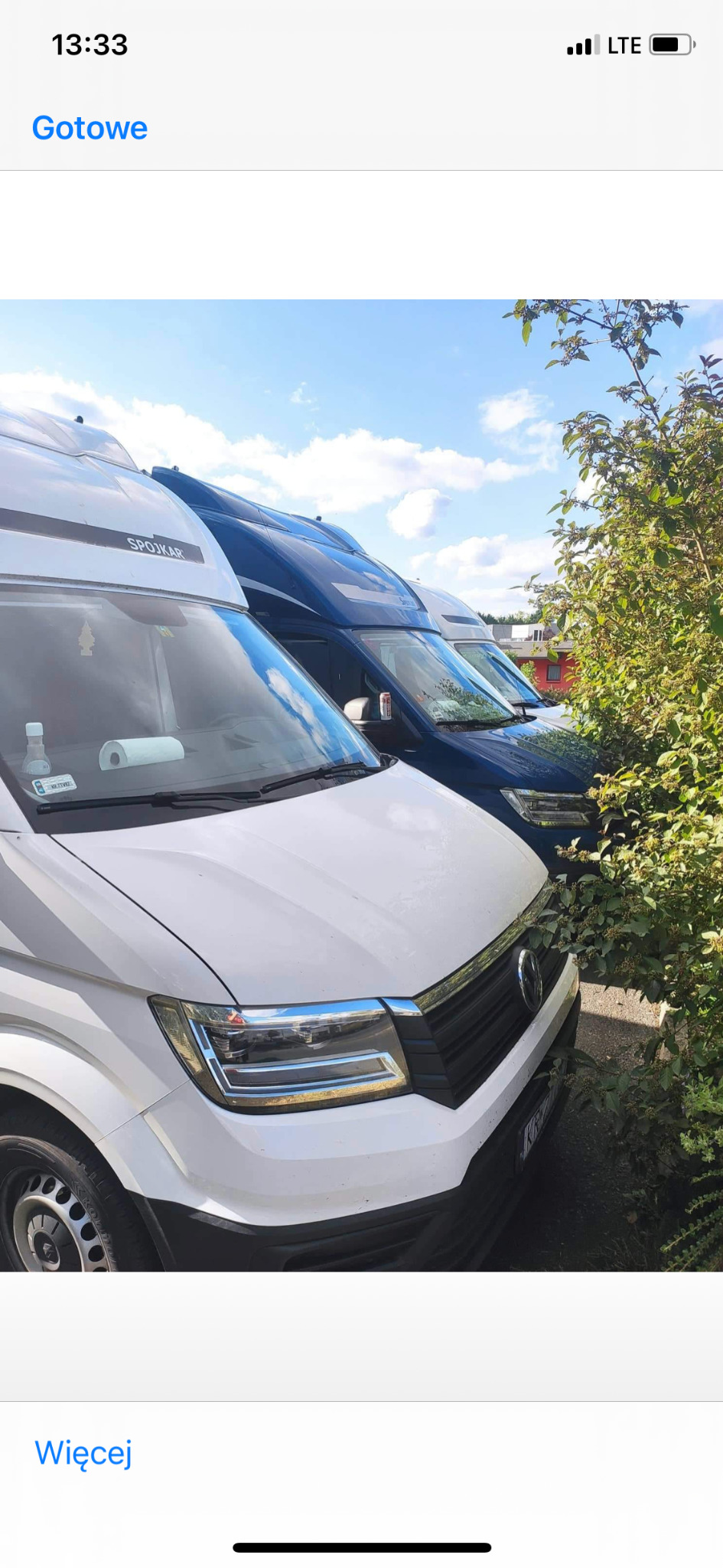 Rząd zaparkowanych busów transportowych, pierwszy biały z widoczną marką SPOJKAR, obok ciemnoniebieski, częściowo zasłonięte przez krzewy