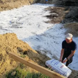 Przygotowanie podłoża pod fundamenty domu: pracownik rozsypuje wapno hydratyzowane na wykopie, w tle drugi pracownik wyrównuje teren.