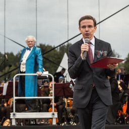 Fotograf - Mężczyzna w garniturze przemawia do mikrofonu na tle orkiestry i drugiego mężczyzny w niebieskiej marynarce.
