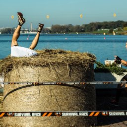 Fotograf - Uczestnik biegu z przeszkodami w białych spodenkach i sportowych butach, z nogami do góry, zanurzony w stogu siana; drugi uczestnik w czarnym stroju sportowym obserwuje z boku, a w tle woda...