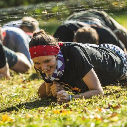 Fotograf - Uśmiechnięta kobieta w czerwonej bandanie czołga się pod drutem kolczastym podczas ekstremalnego biegu przeszkodowego na trawiastym terenie, z innymi uczestnikami w tle.