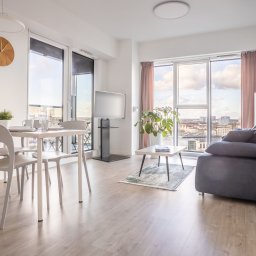 Fotograf - Jasne, nowoczesne wnętrze apartamentu z widokiem na miasto, obejmujące stół jadalny z krzesłami, telewizor na stojaku, sofę z poduszkami i stolik kawowy z rośliną.