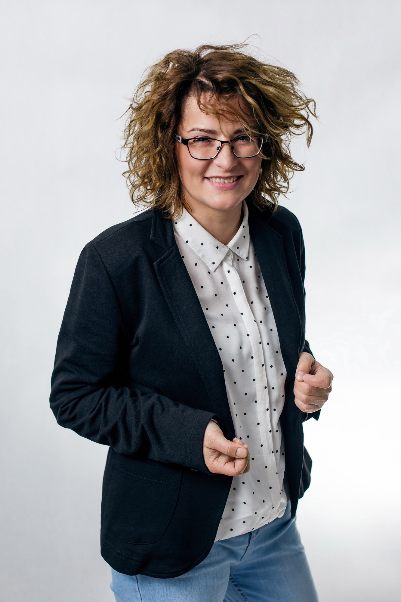 Uśmiechnięta kobieta w okularach, białej koszuli w czarne kropki i czarnej marynarce pozuje na jasnym tle.