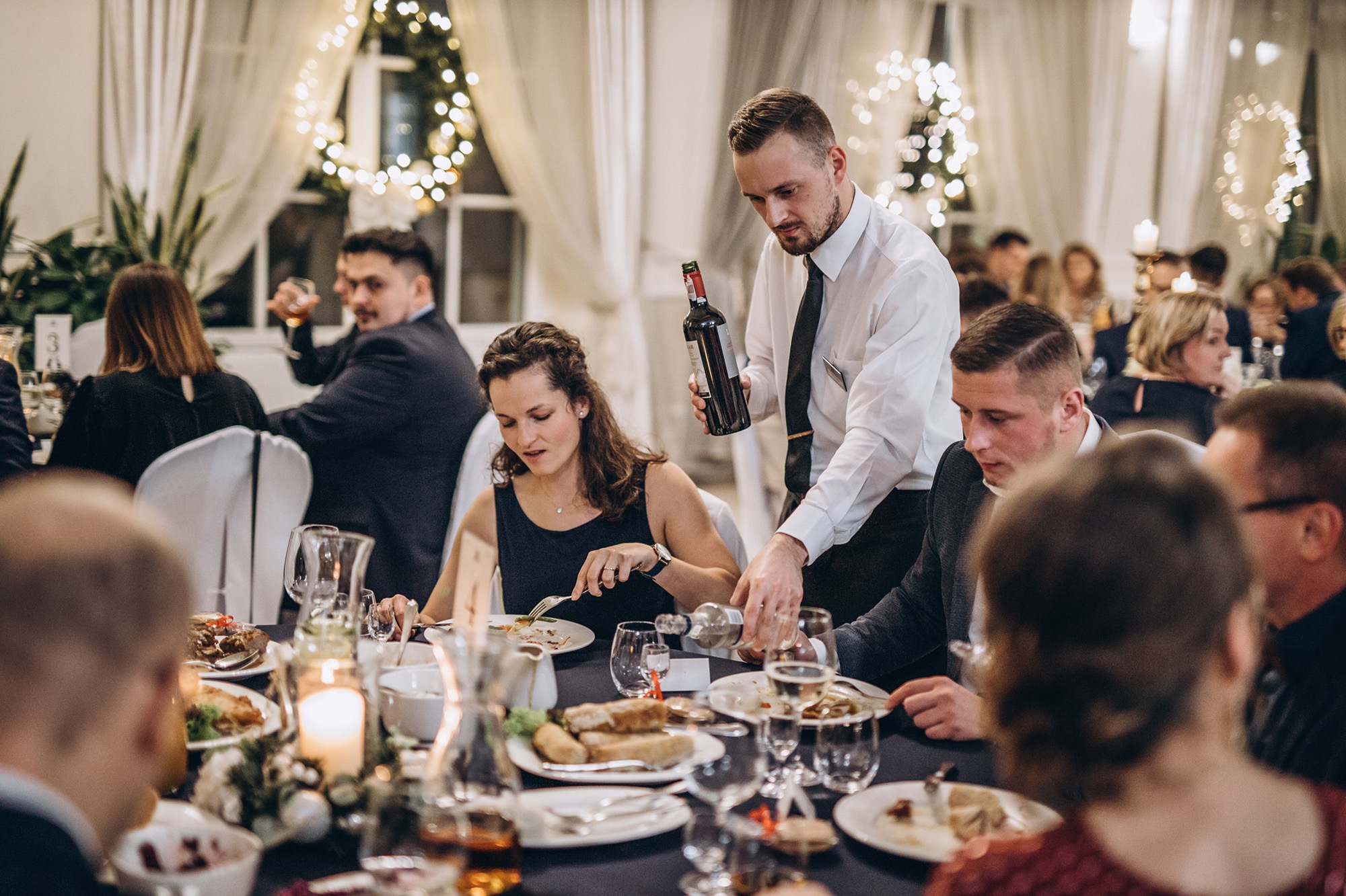 Kelner nalewa wino gościom siedzącym przy stole podczas uroczystości; w tle dekoracje świetlne i okna z białymi zasłonami.