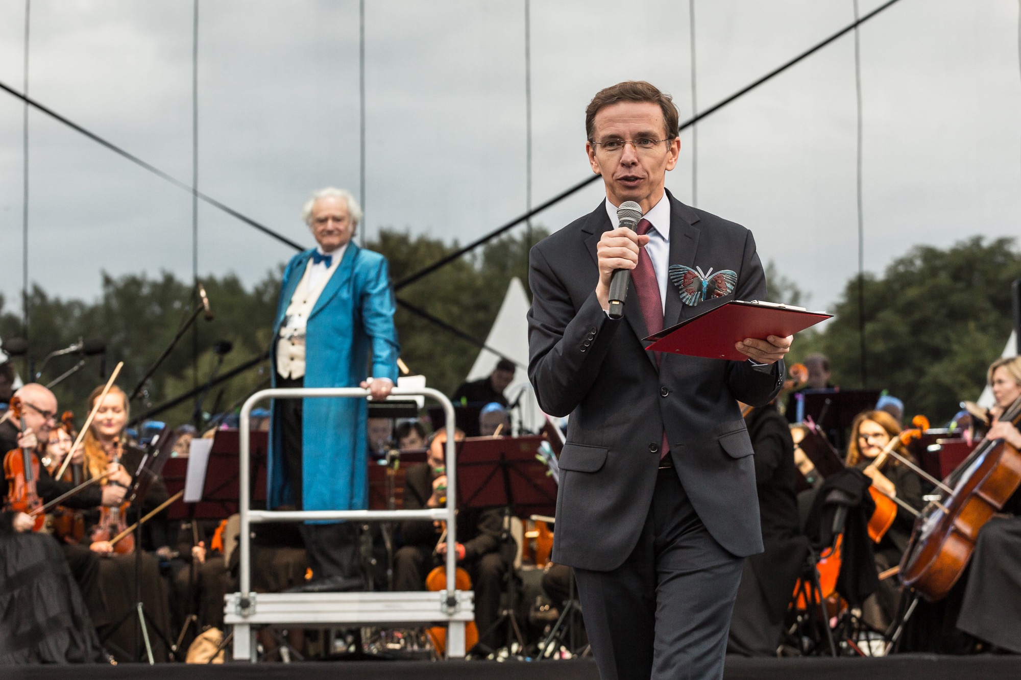 Mężczyzna w garniturze przemawia do mikrofonu na tle orkiestry i drugiego mężczyzny w niebieskiej marynarce.