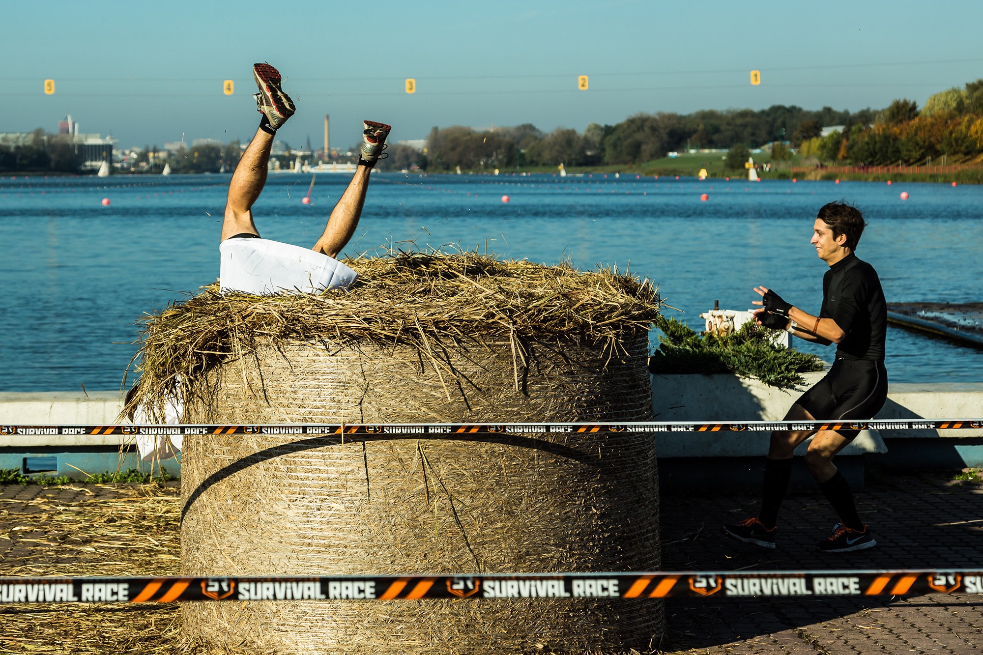 Uczestnik biegu z przeszkodami w białych spodenkach i sportowych butach, z nogami do góry, zanurzony w stogu siana; drugi uczestnik w czarnym stroju sportowym obserwuje z boku, a w tle woda...