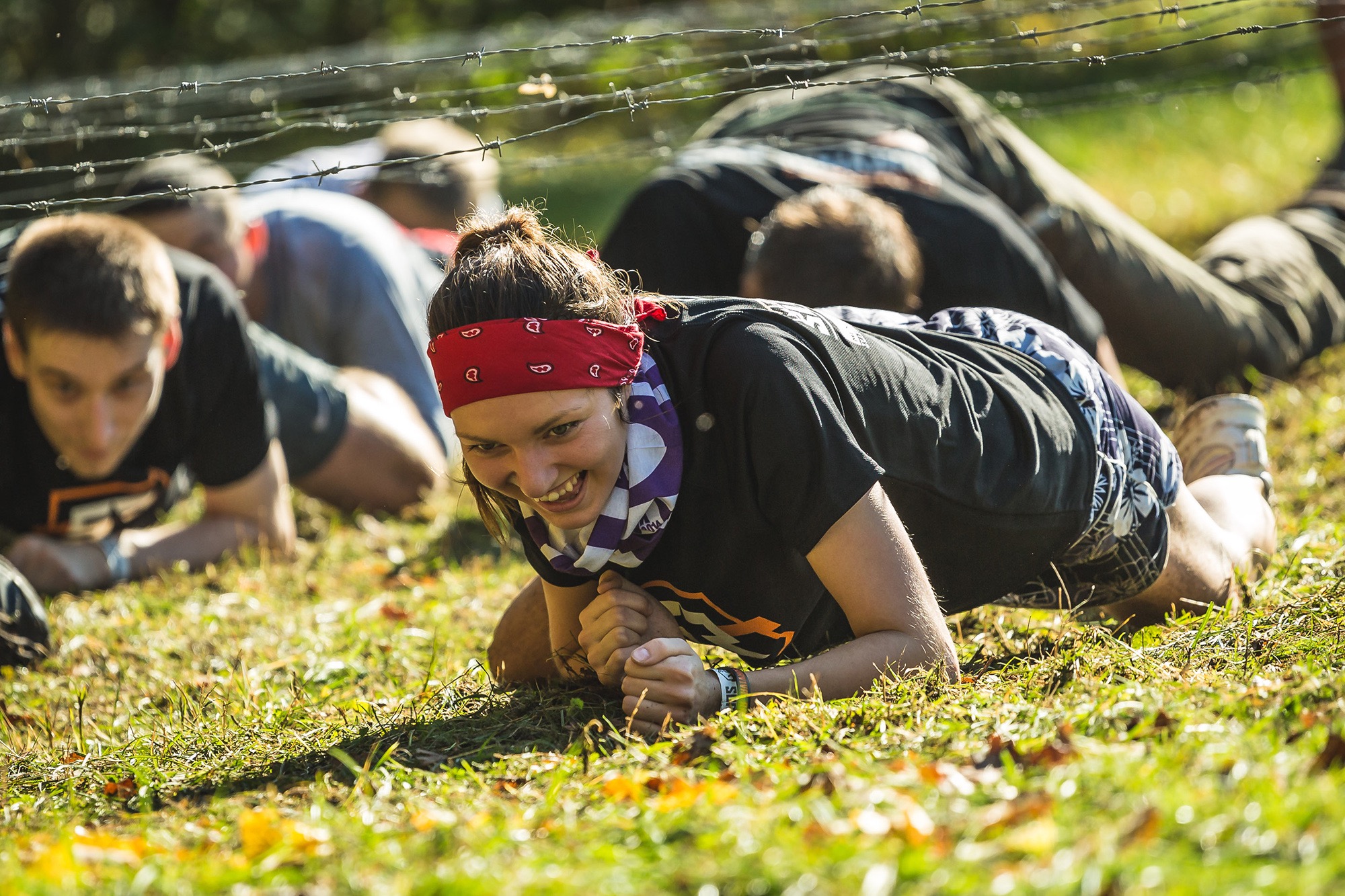 Uśmiechnięta kobieta w czerwonej bandanie czołga się pod drutem kolczastym podczas ekstremalnego biegu przeszkodowego na trawiastym terenie, z innymi uczestnikami w tle.