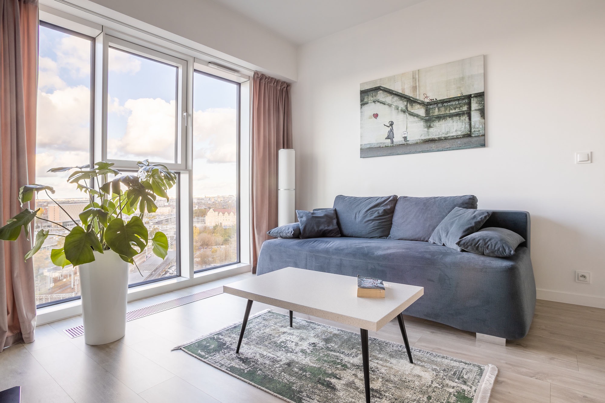 Jasne wnętrze mieszkania z dużym oknem, szarą sofą, stolikiem kawowym i rośliną doniczkową, na ścianie obraz Banksy'ego.