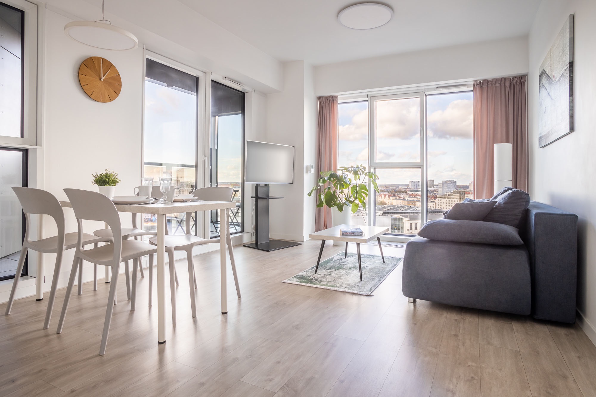 Jasne, nowoczesne wnętrze apartamentu z widokiem na miasto, obejmujące stół jadalny z krzesłami, telewizor na stojaku, sofę z poduszkami i stolik kawowy z rośliną.
