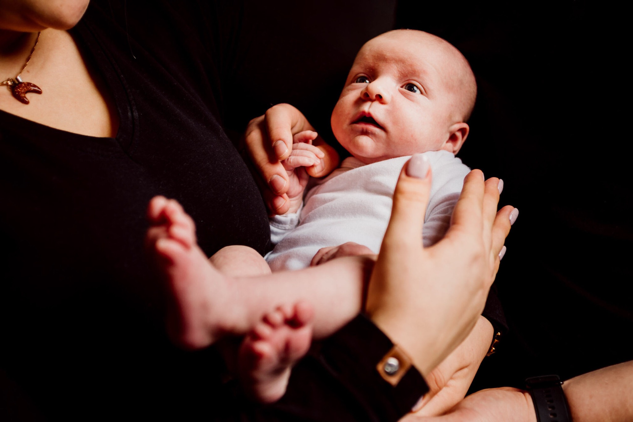 Niemowlę w białym body, trzymane na rękach przez dwie osoby, z widocznym fragmentem czarnej bluzki matki z naszyjnikiem w kształcie muszelki.