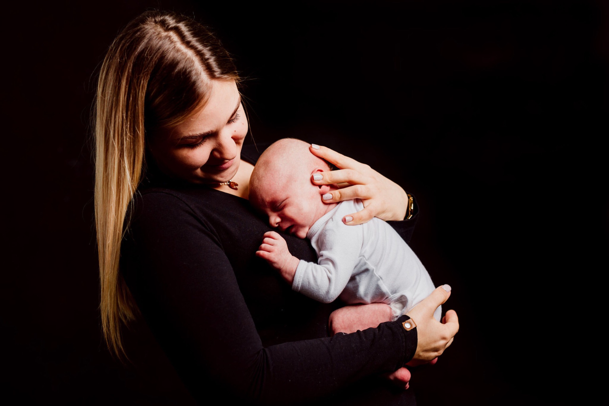 Młoda kobieta z blond włosami trzyma na rękach noworodka w białym body na czarnym tle, delikatnie się uśmiechając.