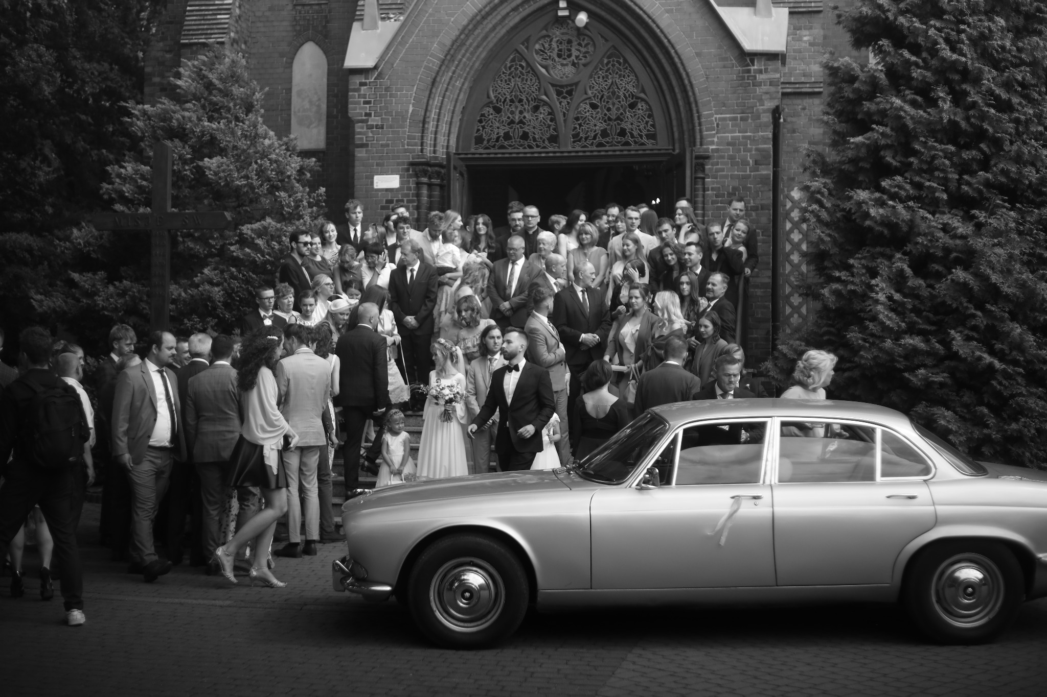 Czarno-białe zdjęcie nowożeńców i gości weselnych wychodzących z kościoła, z klasycznym srebrnym samochodem na pierwszym planie.