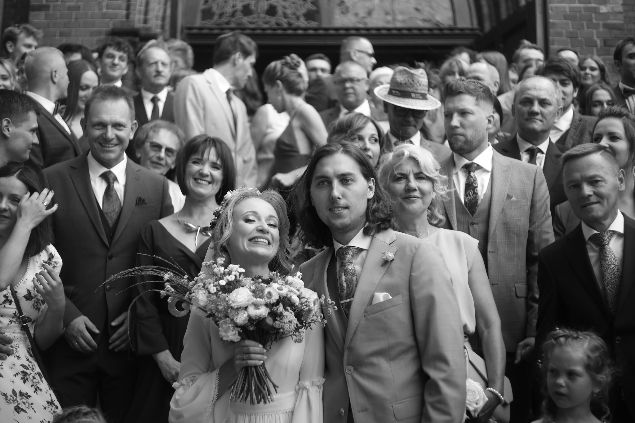 Czarno-białe zdjęcie uśmiechniętej panny młodej z bukietem i pana młodego na tle grupy gości weselnych przed budynkiem z cegły.
