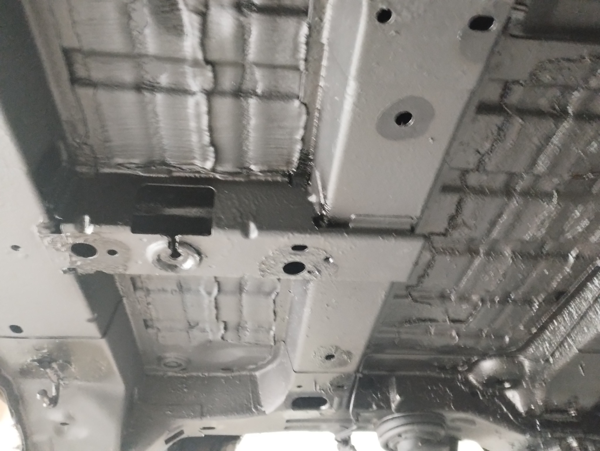 Podwozie samochodu po nałożeniu warstwy ochronnej antykorozyjnej, widoczne liczne otwory montażowe i elementy konstrukcyjne pokryte grubą, szarą warstwą preparatu.