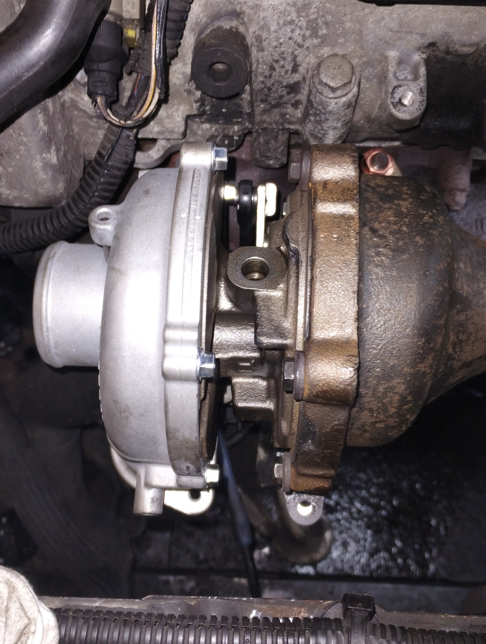 Turbosprężarka silnika samochodowego, widoczne ślady użytkowania i zabrudzenia, nowa część zamontowana, metalowe elementy złączone śrubami.