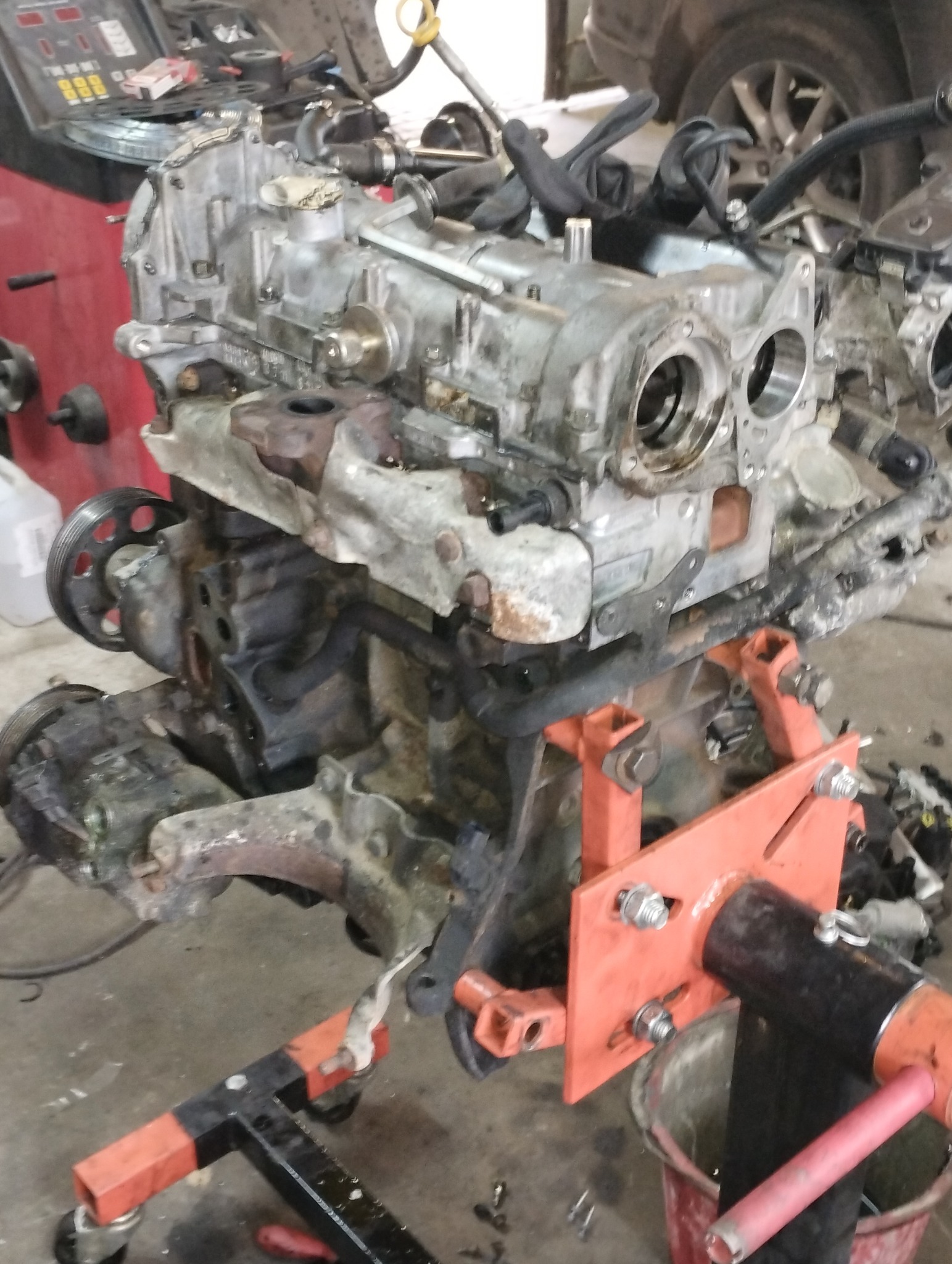 Zdemontowany, brudny silnik samochodowy na pomarańczowym stojaku w warsztacie, widoczne elementy osprzętu i przewody.