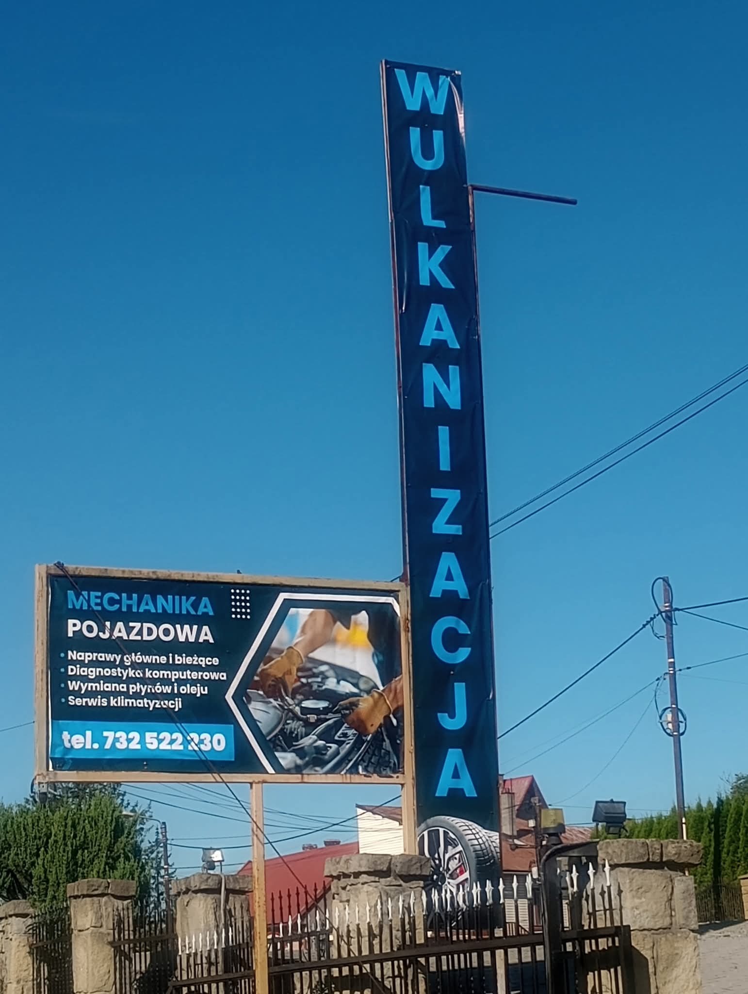 Reklamowy szyld warsztatu samochodowego z napisem 'Wulkanizacja' i ofertą usług mechanicznych, w tle błękitne niebo i fragment ogrodzenia.