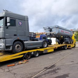 PSP TRANS SPÓŁKA Z OGRANICZONĄ ODPOWIEDZIALNOŚCIĄ - Szary ciągnik DAF XF 105.510 z logo na kabinie transportowany na żółtej niskopodwoziowej platformie lawety wraz z naczepą MS Transporte na tle pochmurnego nieba i miejskiej infrastruktury.