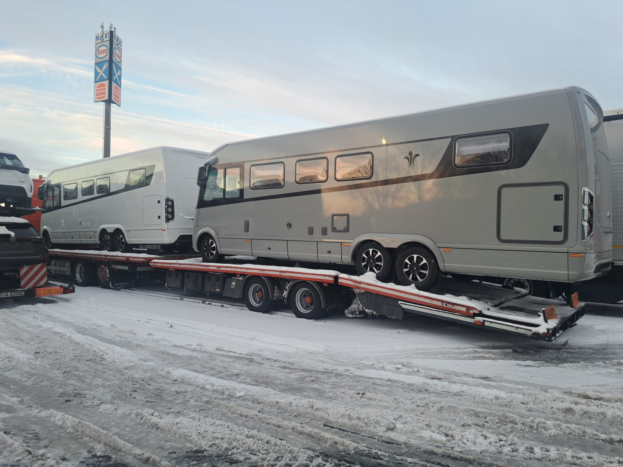 Dwa luksusowe kampery na niskopodwoziowej platformie transportowej, widoczne na tle zasnieżonego parkingu z dystrybutorem paliw i fast foodem, wskazują na transport specjalistyczny.