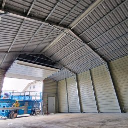 Bartosz Wójtowicz Biuro Projektowe - Wnętrze hali stalowej w trakcie montażu, widoczny pracownik na podnośniku koszowym i konstrukcja dachu z blachy trapezowej.