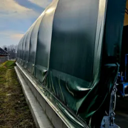 Zielona, plandekowa hala namiotowa z metalową konstrukcją, widok z zewnątrz, w tle błękitne niebo z chmurami.