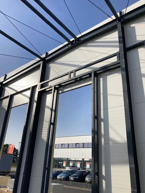 Konstrukcja hali w trakcie budowy, widoczne stalowe ramy i białe panele ścienne, na zewnątrz zaparkowane samochody i budynek biurowy w tle.