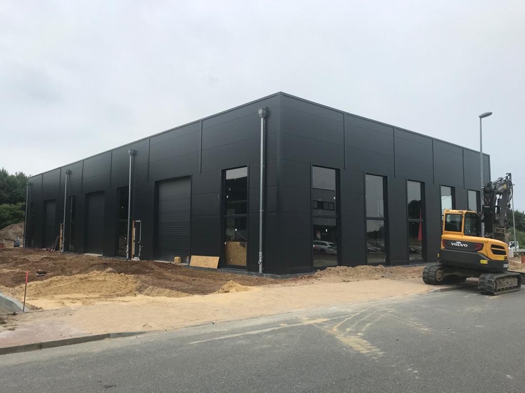 Nowoczesna hala biurowo-magazynowa o czarnej, modułowej elewacji z widocznymi oknami i bramami rolowanymi, obok żółta koparka Volvo na placu budowy.