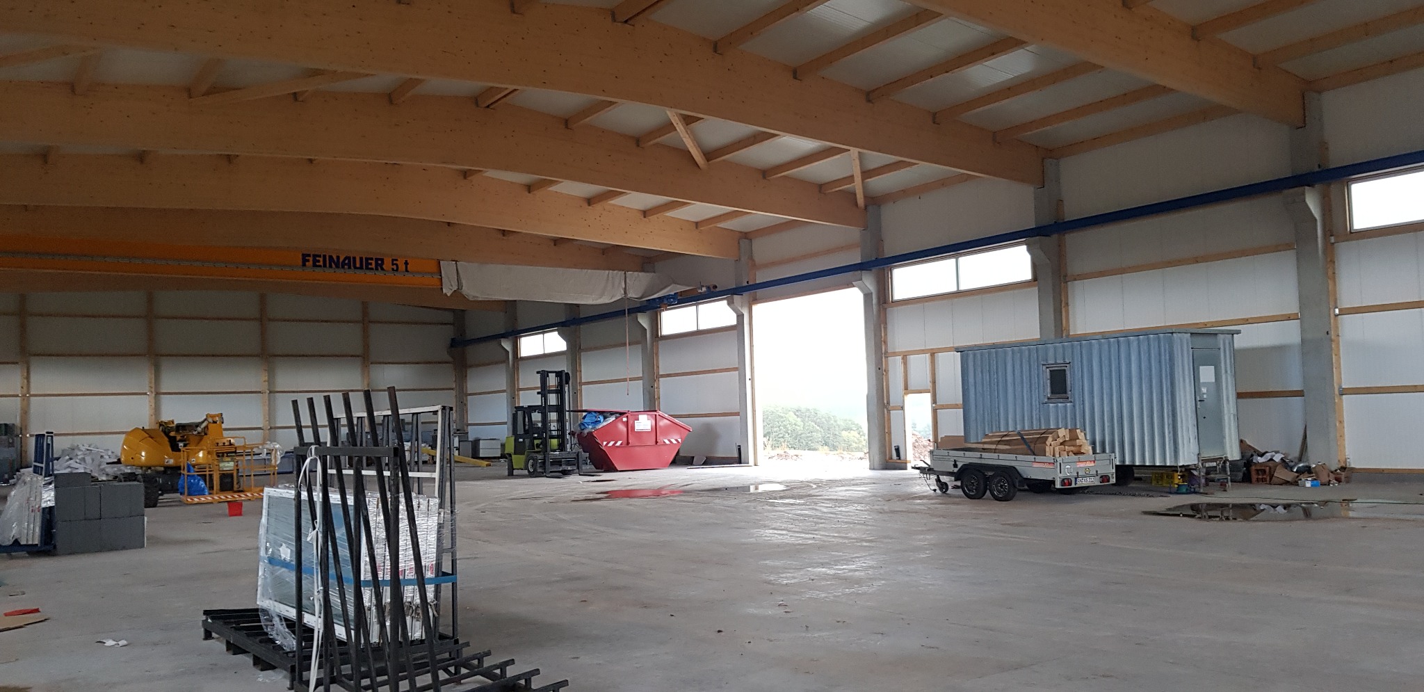 Wnętrze hali z drewnianą konstrukcją stropu, widoczny suwnica, wózki widłowe, kontener, przyczepa z drewnem oraz okna doświetlające przestrzeń.