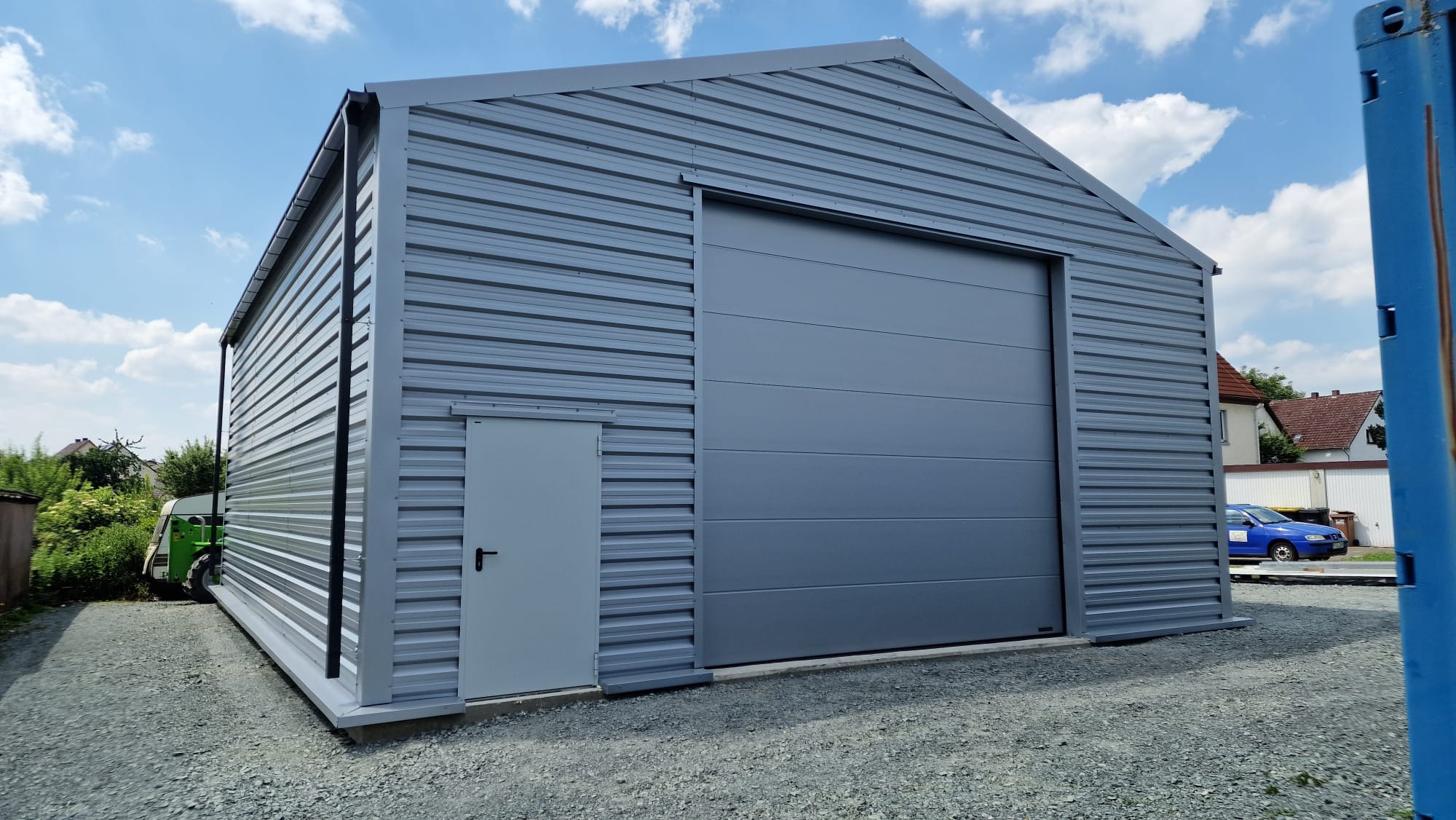 Szara hala z blachy trapezowej z bramą przesuwną i drzwiami wejściowymi, widok z zewnątrz na tle błękitnego nieba z chmurami.