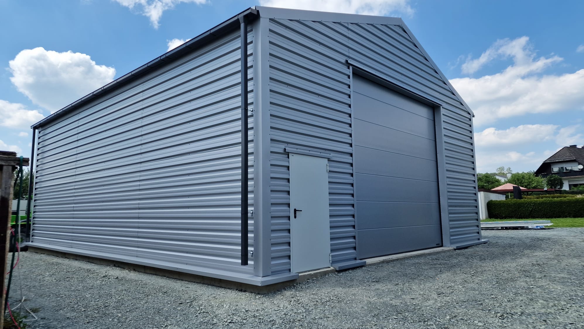 Gotowa hala stalowa w kolorze szarym z bramą segmentową i drzwiami wejściowymi, widoczna na tle błękitnego nieba z chmurami.